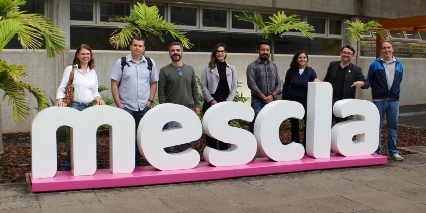 Foto de 8 pessoas posicionadas atrás de um letreiro branco com detalhe rosa escrito "Mescla". Fim da descrição.
