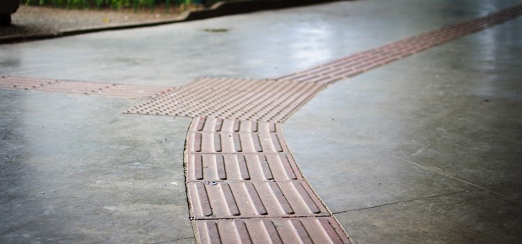 Foto de calçada de cimento acinzentado com trilha de piso tátil para pessoas com deficiência visual. Fim da descrição.