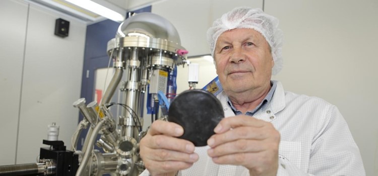 Foto colorida do professor Stanislav segurando uma amostra da borracha, um circulo preto. Ao fundo, há uma torre prateada que faz parte de um dos equipamentos do laboratório. Fim da descrição.
