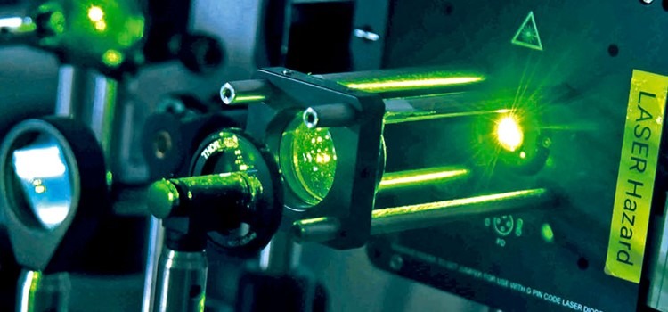 Foto colorida focada em parte de um microscópio que emite luz verde. Fim da descrição.