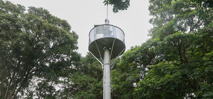 Foto de torre metálica em ambiente externo e arborizado. Fim da descrição.