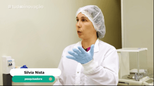 Print do vídeo mostra a pesquisadora Silvia Nista com jaleco branco e touca, em ambiente de laboratório. Fim da descrição.