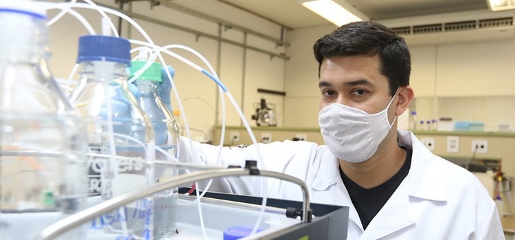Foto do professor Daniel em ambiente de laboratório. Ele veste máscara e mexe em equipamento com fios e garrafas interconectadas. Fim da descrição.