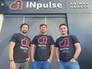 Foto mostra três homens brancos em frente a fachada da empresa INpulse. Eles veste camiseta preta com o logo da empresa em vermelho no formato de um coração. Fim da descrição.