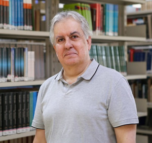 Homem branco de cabelos grisalhos, veste camisa polo cinza e está na frente de prateleiras de livros.
