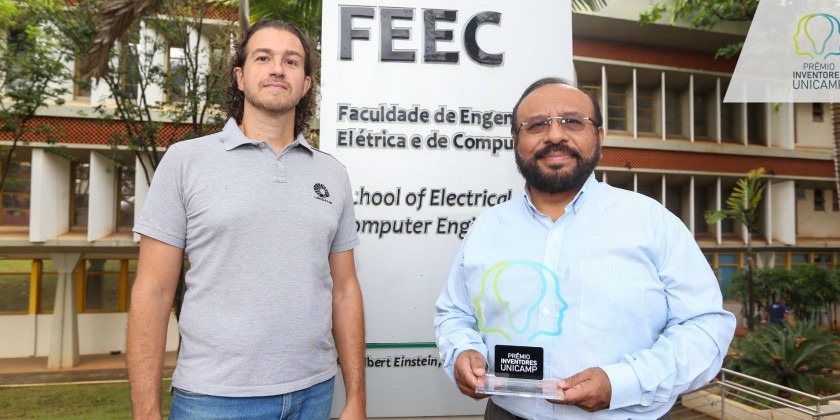 Homem de camisa cinza (a esquerda) e homem de branco (a direita) segurando a premiação em frente a placa da FEEC