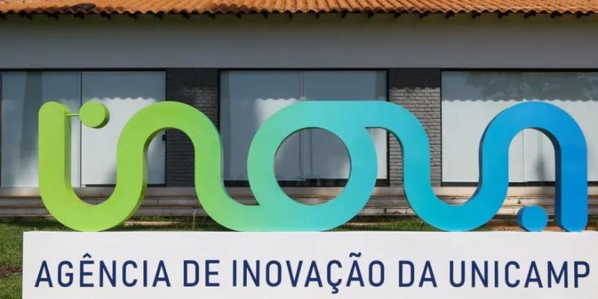 A imagem mostra uma placa da “Agência de Inovação da UNICAMP” no Brasil. A placa é composta pela palavra “Inova” em uma fonte estilizada, com as letras “I” e “n” em verde e as letras “o”, “v” e “a” em azul. A placa está sobre uma base de concreto branco com as palavras “Agência de Inovação da UNICAMP” em letras pretas. O fundo consiste em um prédio com telhado vermelho e paredes brancas.