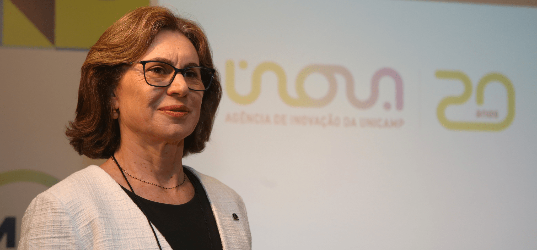 Na foto está a professora Ana Frattini, uma mulher branca, com cabelos castanhos na altura do pescoço usando óculos de grau. A foto é colorida e ela está a frente de um banner com a projeção do logotipo da Inova Unicamp. Fim da descrição.