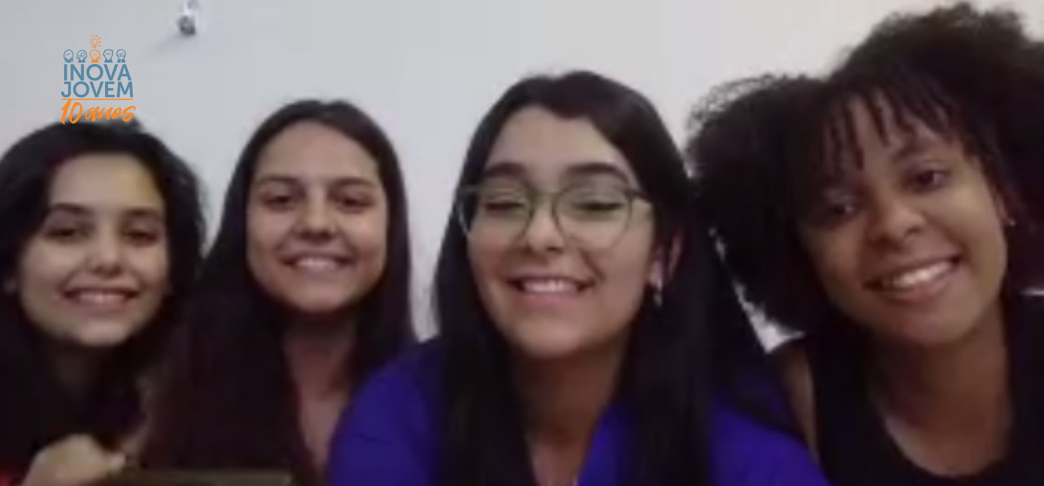Captura de tela. Quatro meninas, três brancas e uma negra sorriem para a câmera.