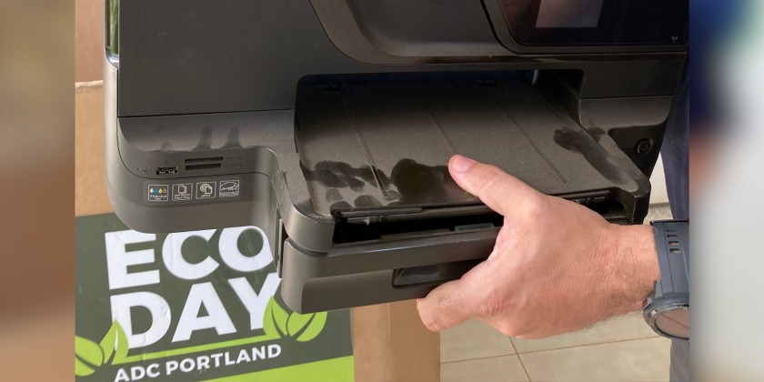 Ação de descarte consciente no Eco Day: Uma mão está colocando uma impressora preta antiga em uma lixeira de reciclagem com um rótulo verde que diz ‘Eco Day’. O fundo é uma parede bege com uma placa verde que diz ‘ADC Portland’. A face e o corpo da pessoa estão desfocados, apenas a mão é visível.