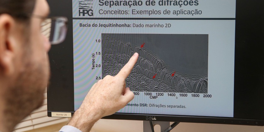 Fotografia. Homem aponta para tela de computador onde está escrito: Separação de Difrações, Conceitos: exemplos de Aplicação e abaixo um gráfico. Fim da descrição.