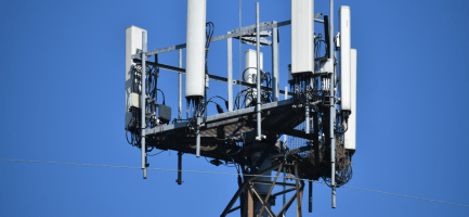 Imagem externa retrata uma torre de celular. Nela, há um céu azul ao fundo e um equipamento alto com estrutura de transmissão de sinal. Fim da descrição.