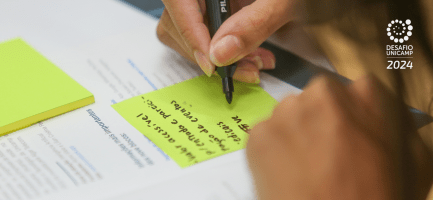 A imagem é uma fotografia em que aparece uma mão escrevendo com uma caneta preta em um post-it. O post-it está em cima de um modelo de negócios impresso. A imagem está ilustrando a atividade de modelagem de negócios do Desafio Unicamp 2024. Fim da descrição.