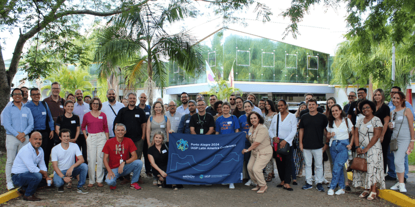 Foto posada dos participantes da Conferência IASP América Latina. Imagem feita em ambiente externo conta com diversas pessoas, em posições diferentes, com algumas segurando a bandeira da IASP. Fim da descrição.