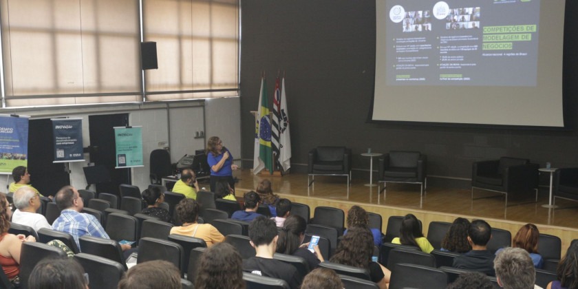 Foto colorida de auditório onde pessoas assistem à apresentação da Inova Unicamp. Iara Ferreira, mulher branca, apresenta dados que são projetados em um telão. Fim da descrição.