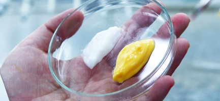 Foto em close mostra a mão de uma pessoa segurando um recipiente de vidro onde há duas soluções em textura de creme posicionadas lado a lado. Uma é amarela e a outra branca. Fim da descrição.