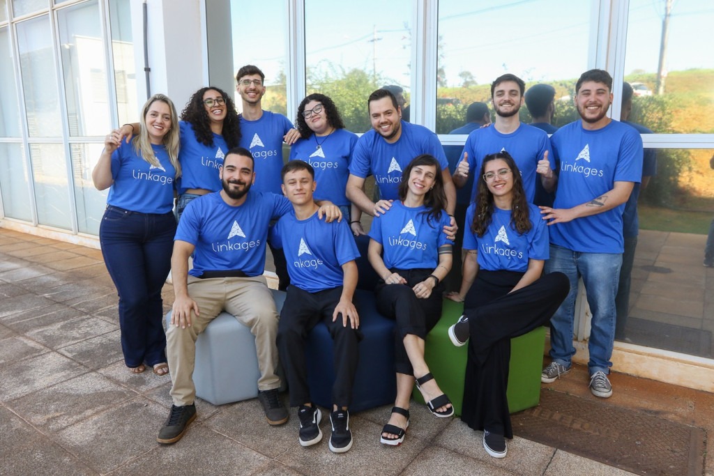 Fotografia de grupo de funcionários da linkages em área externa, todos usam camisetas azuis da empresa, alguns se encontram sentados enquanto outros, atrás, de pé. Fim da descrição.