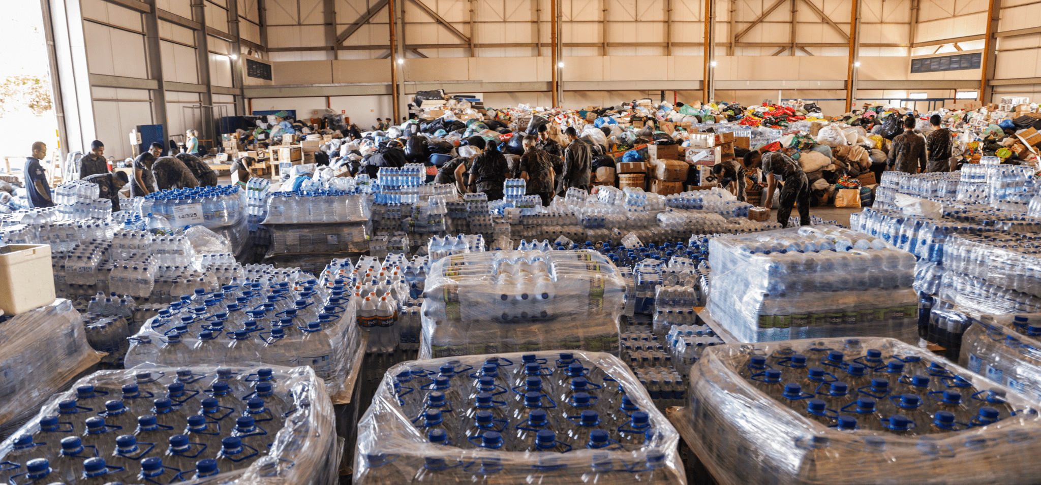 Foto da base aérea de Brasília de recebimento de doações. Registro conta com diversas garrafas d'água à frente do plano, com caixas de alimentos ao fundo. Fim da descrição.