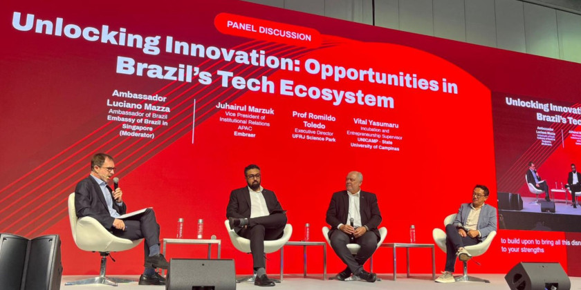 Foto colorida mostra quatro homens sentados em uma poltrona e um telão de apresentações vermelho ao fundo. Fim da descrição.